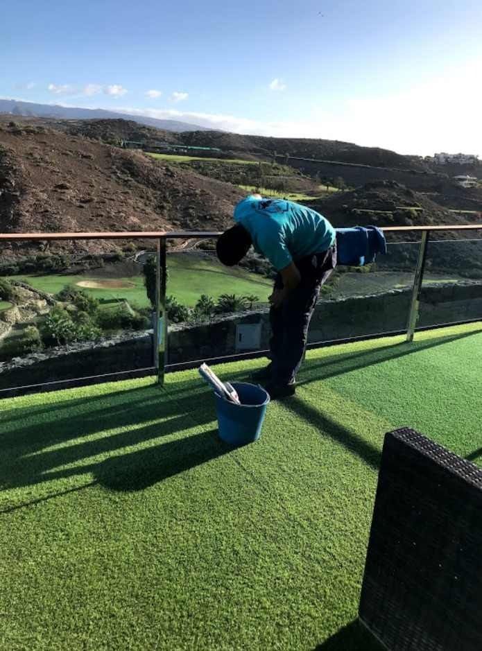 Persona limpiando una barandilla de vidrio en un patio con césped verde con vista a un campo de golf y colinas en un día soleado.