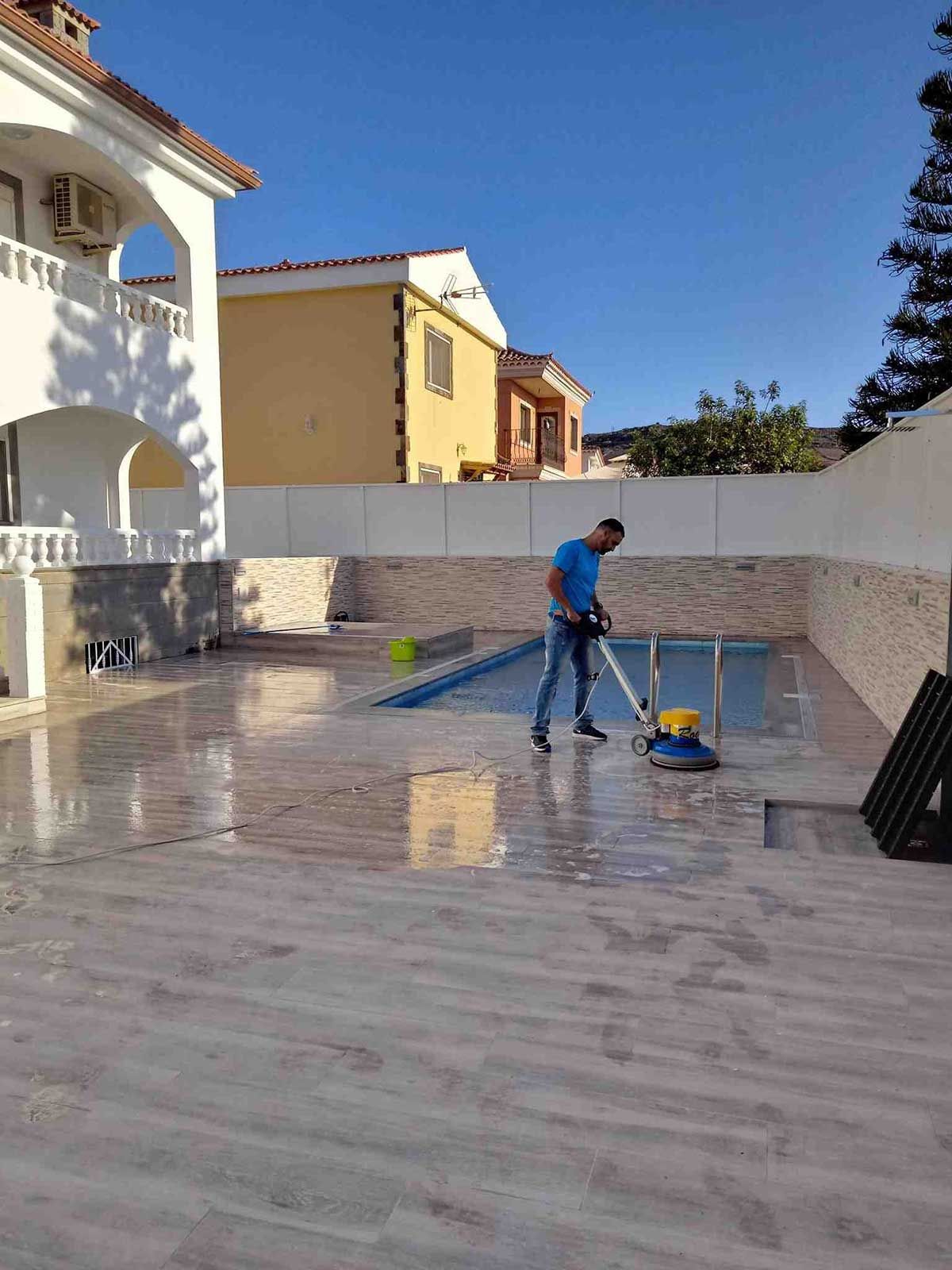 Hombre limpiando el patio alrededor de una piscina con una máquina; casas y cielo azul en el fondo.