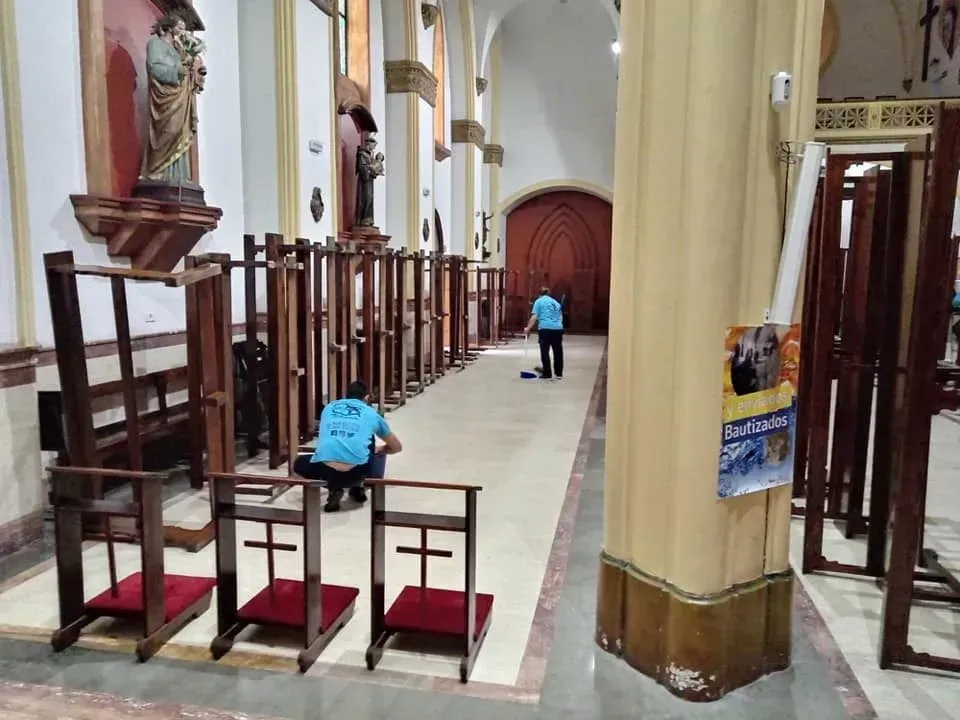 Personas limpiando una iglesia con filas de reclinatorios de madera.