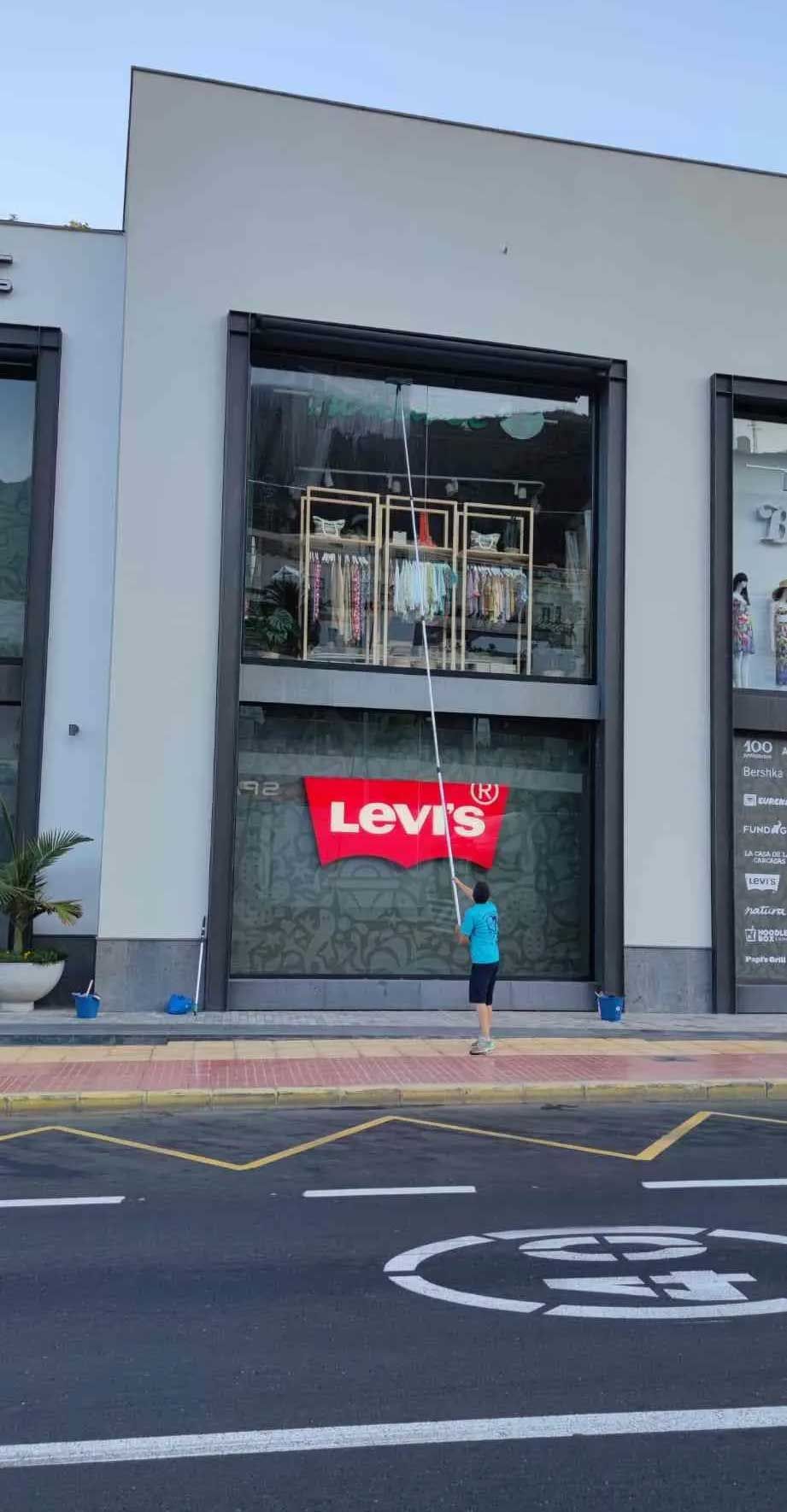 Persona colgando un cartel de Levi's en la ventana de un edificio. Camisa azul, pantalones cortos negros. Edificio blanco, cielo azul.