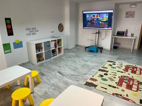Sala de juegos luminosa con juguetes, televisor, mesas, taburetes y alfombras coloridas sobre un suelo gris.