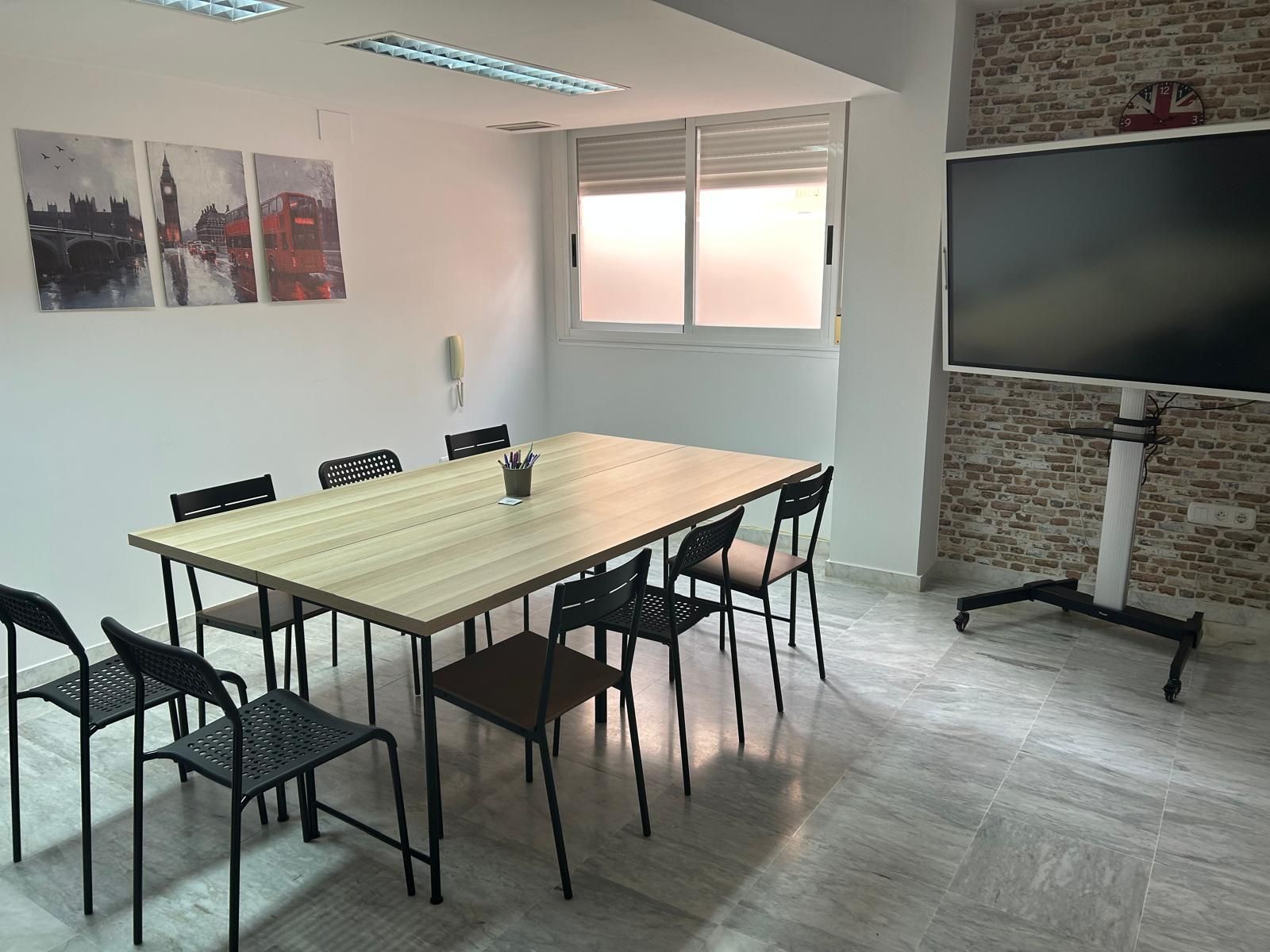 Sala de reuniones moderna y vacía con mesa larga, sillas negras, obras de arte en la pared y un televisor grande en una pared decorativa de ladrillo.