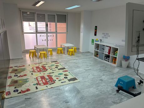 Aula vacía con una alfombra de juegos colorida, sillas amarillas pequeñas, estantes de almacenamiento y una pared con ventana.