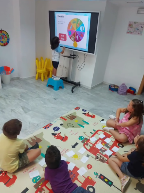 Los niños están sentados en una alfombra del aula viendo una lección en una pantalla montada en la pared.