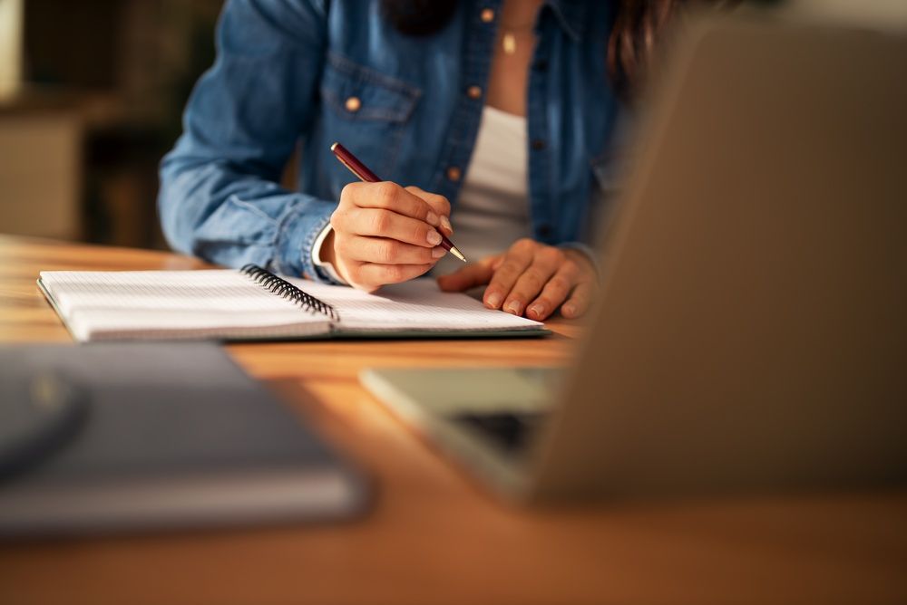 Una persona escribe en un cuaderno en un escritorio con una computadora portátil cerca.