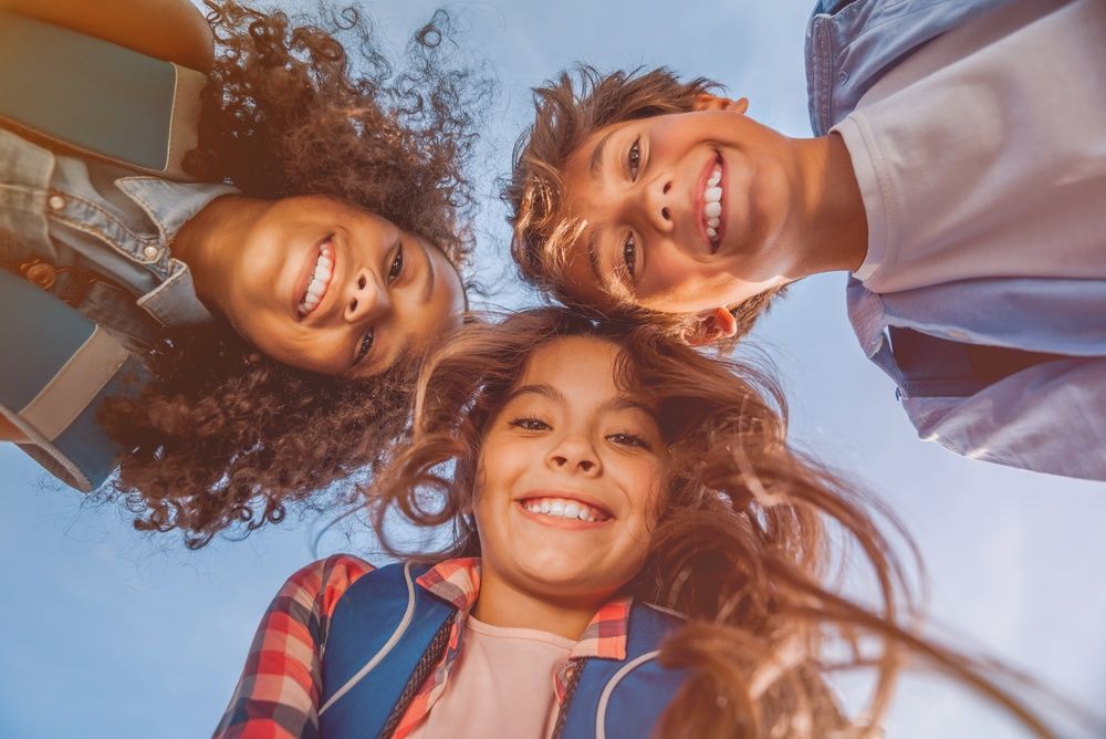 Tres amigas sonrientes, con cabello rizado y chaquetas coloridas, se inclinan sobre la cámara contra un cielo azul.