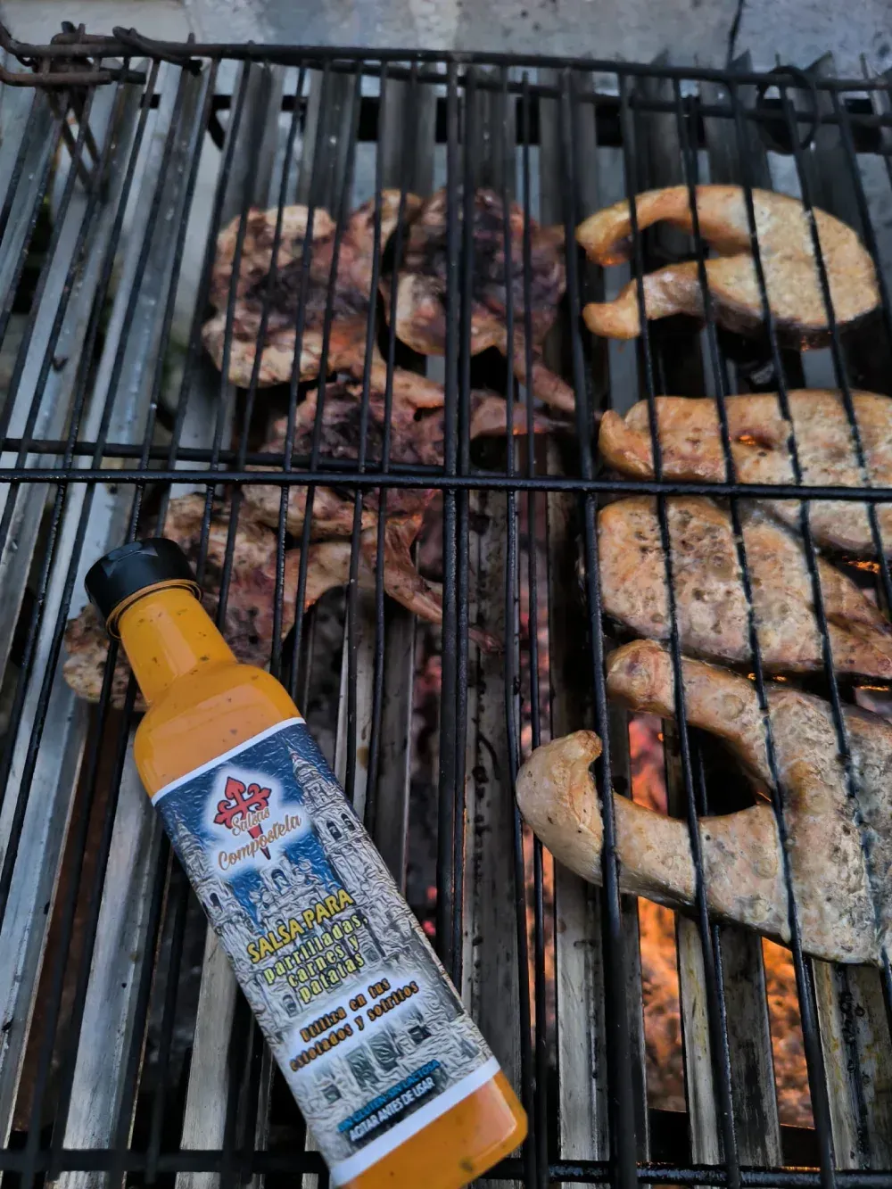 Carne a la parrilla y filetes de salmón sobre una parrilla caliente con una botella de salsa amarilla en primer plano.