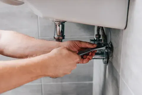 Persona que usa una llave para apretar las tuberías de plomería debajo del lavabo del baño.