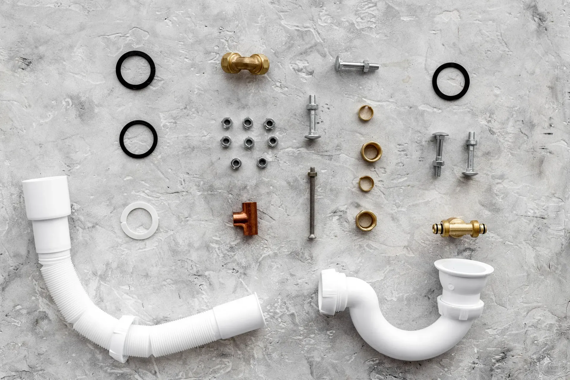 Piezas de fontanería dispuestas sobre una superficie de hormigón gris. Incluye tuberías, conectores, arandelas y tuercas.