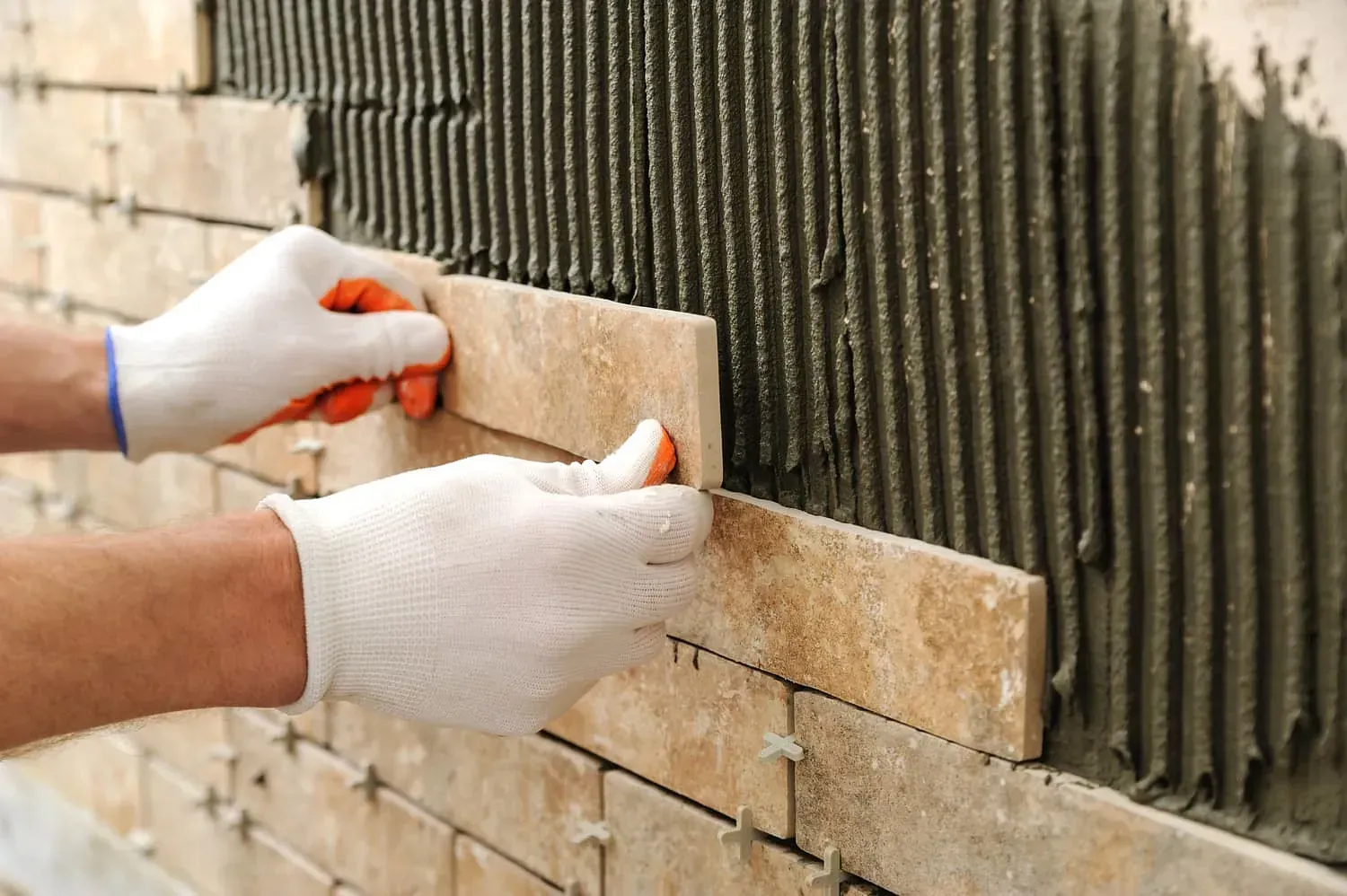 Manos en guantes instalando azulejos beige en una pared cubierta con mortero.
