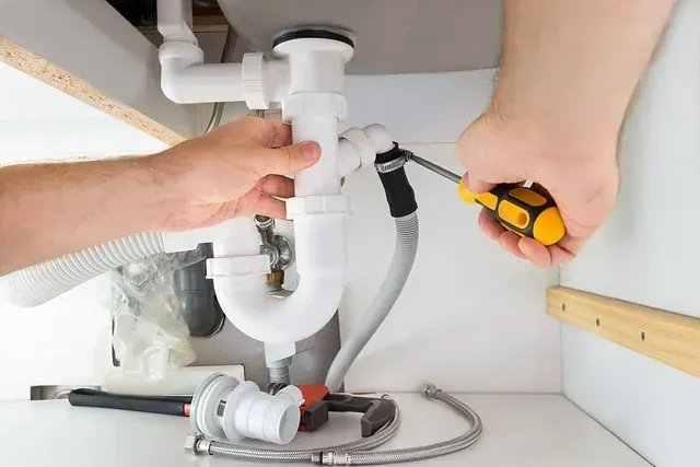 Manos de una persona arreglando una tubería de fregadero blanco debajo de un gabinete blanco, usando un destornillador.