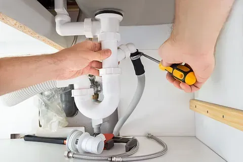 Manos de un fontanero trabajando en las tuberías del fregadero blanco debajo de un armario, utilizando un destornillador.