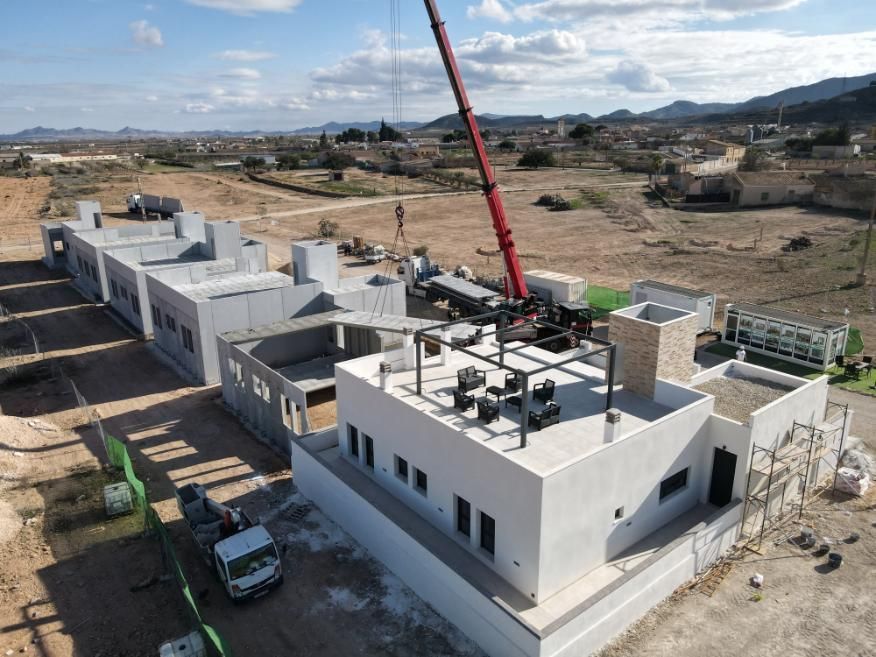 Obra en construcción con edificios blancos, grúa y vehículos en un paisaje seco y abierto.