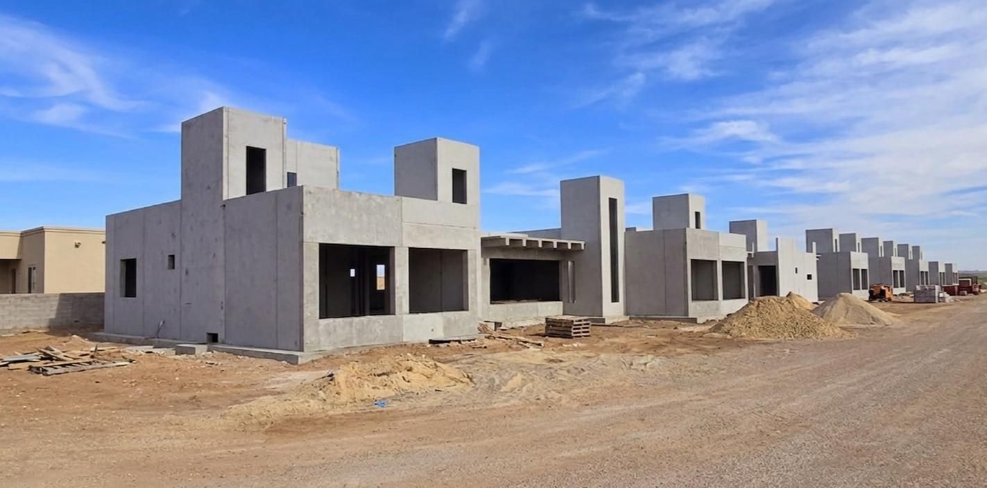 Hilera de casas de hormigón sin terminar en construcción en un paisaje desértico bajo un cielo azul.