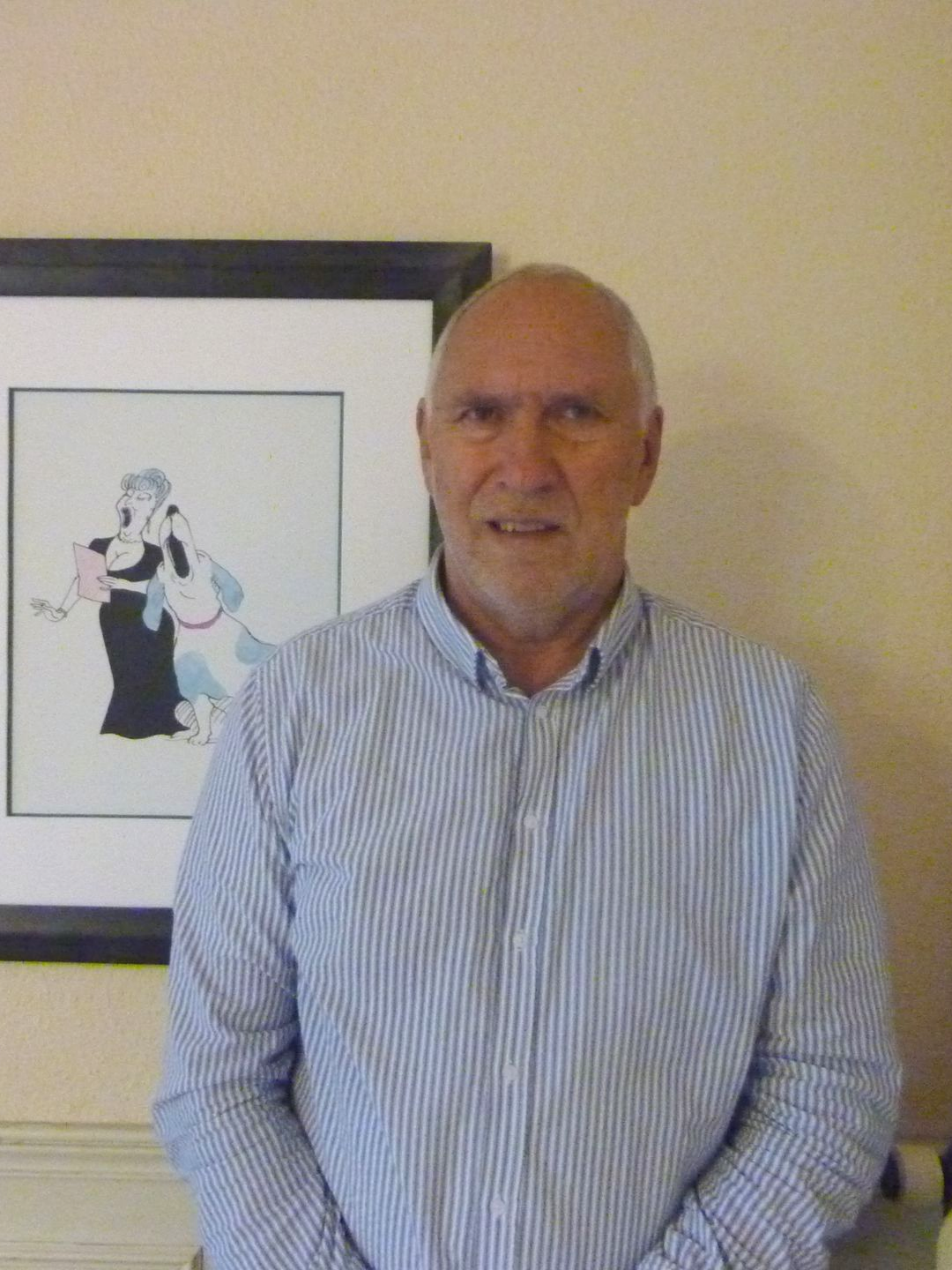 Un homme en chemise à rayures bleues sourit, debout devant un dessin animé encadré.