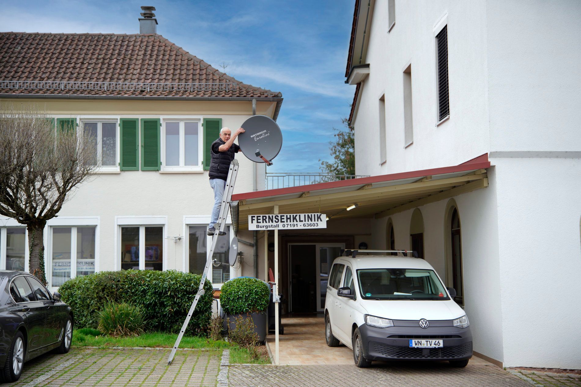 Eine Person montiert auf einer Leiter eine Satellitenschüssel an der Außenwand eines Gebäudes in der Nähe eines Parkplatzes.