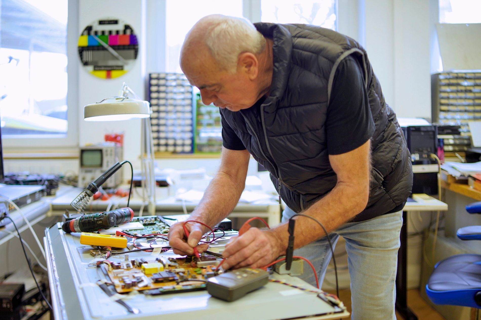 Eine Person in einer Schutzweste testet an einer Werkbank mit Multimeter-Messspitzen elektronische Bauteile auf einer Leiterplatte.