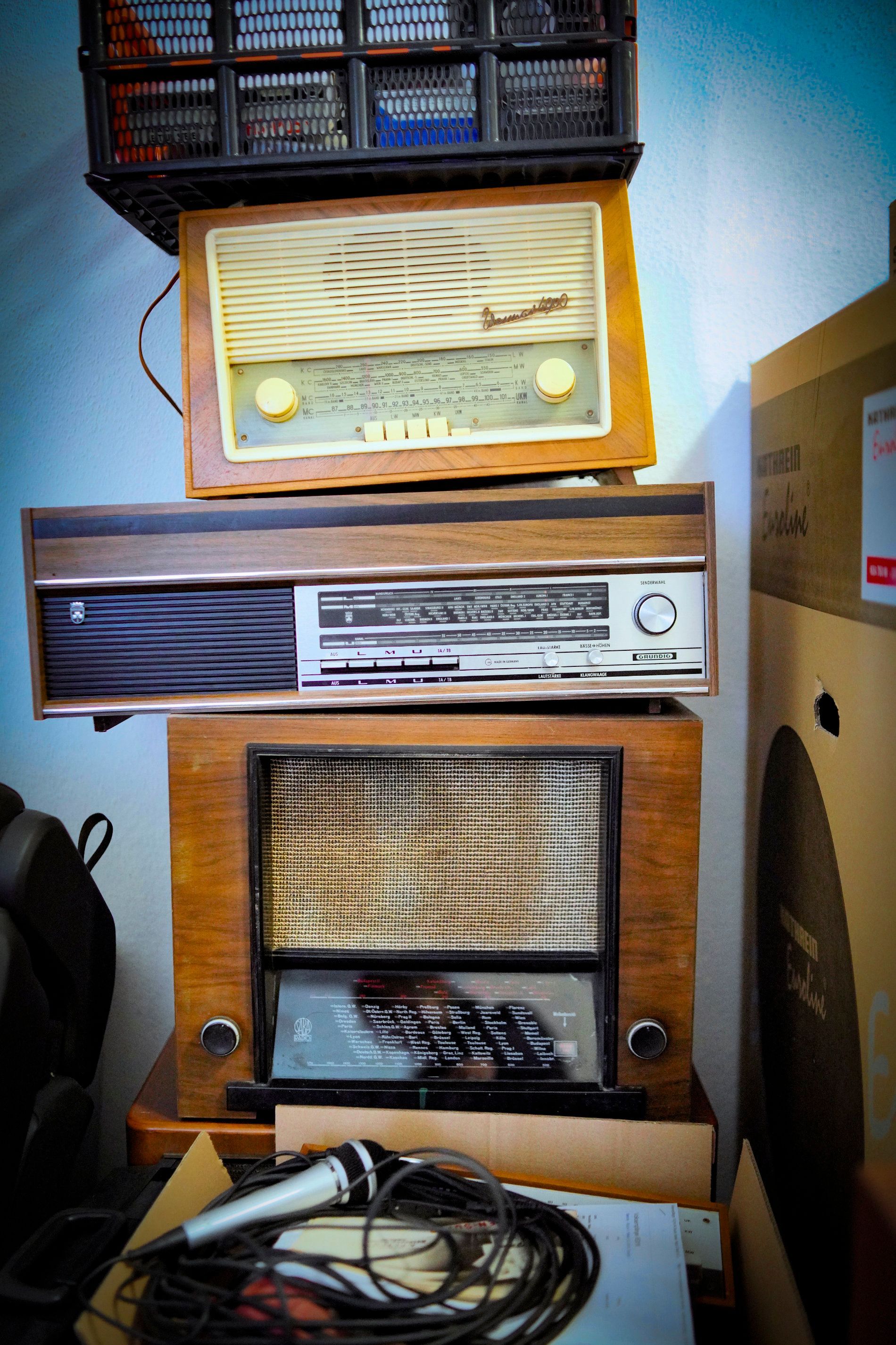 Ein Stapel alter hölzerner Radios und Audiogeräte, im Vordergrund ein aufgewickeltes Mikrofon.