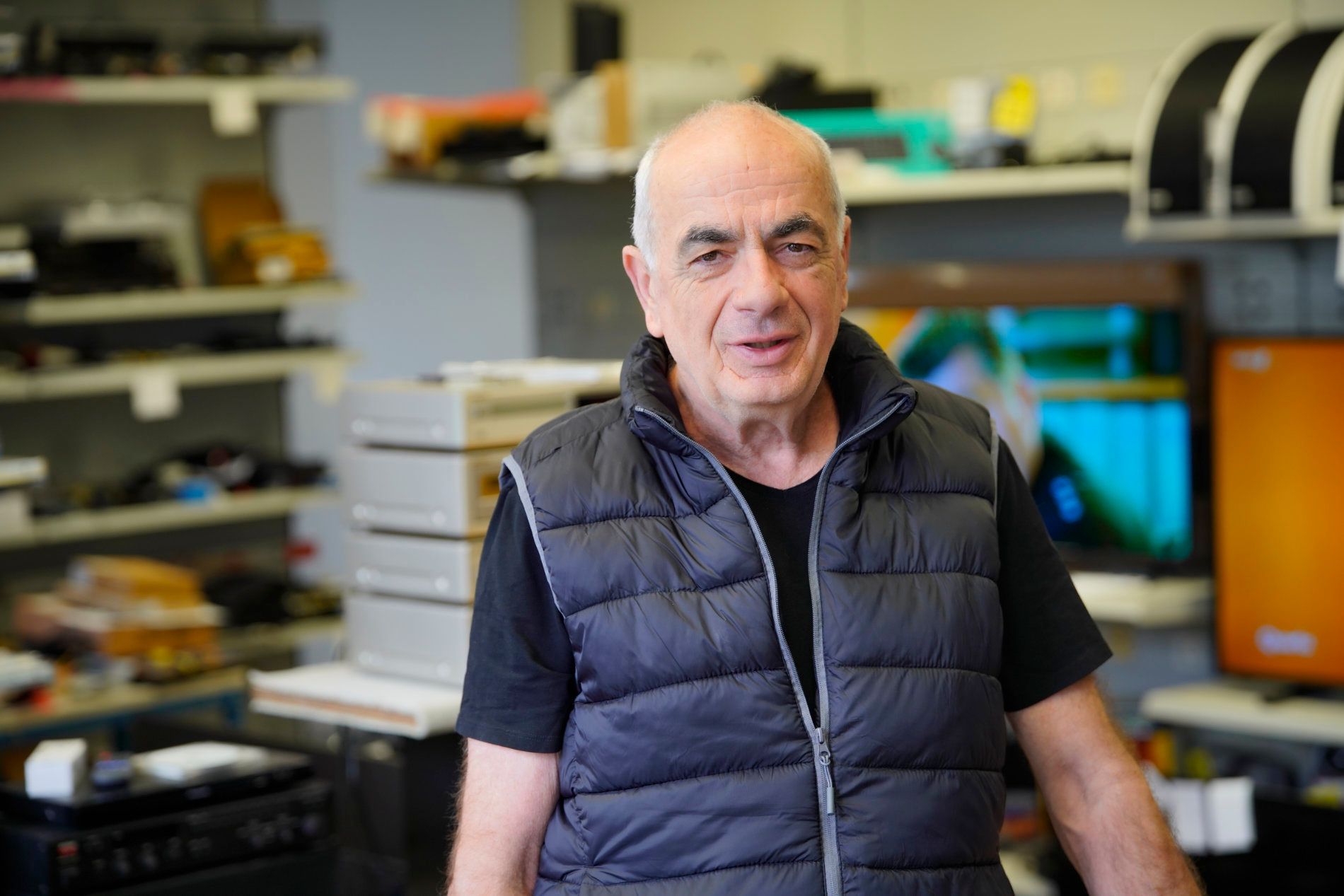 Eine Person steht in einer Werkstatt oder einem Geschäft voller elektronischer Geräte und Regale und blickt in die Kamera.