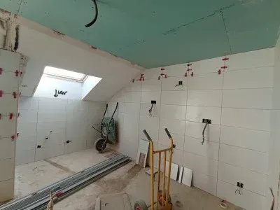 Una habitación en obras con paredes de azulejos blancos, una claraboya, un techo con paneles verdes, una carretilla y una plataforma rodante.