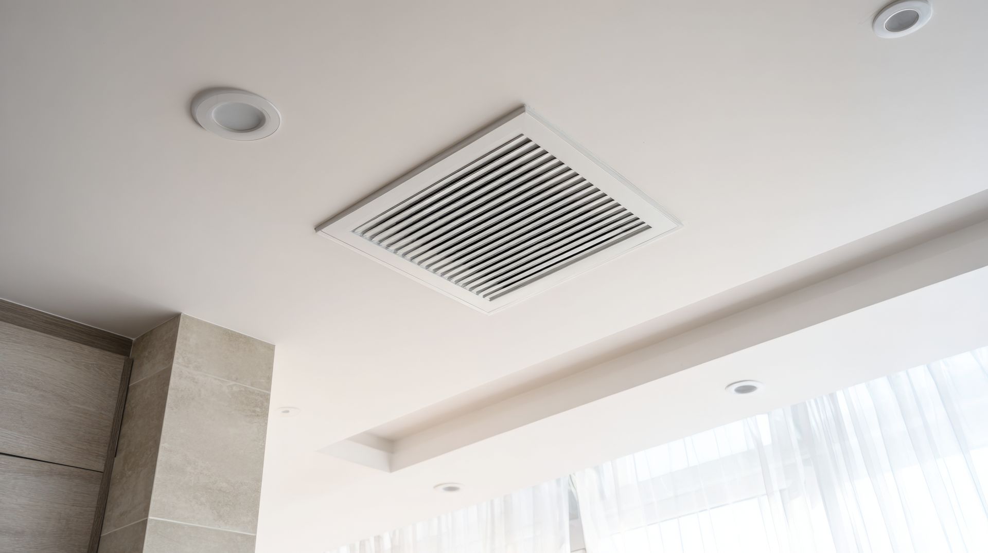 Plafond blanc avec grille d'aération carrée, spots encastrés et fenêtre à voilage en dessous