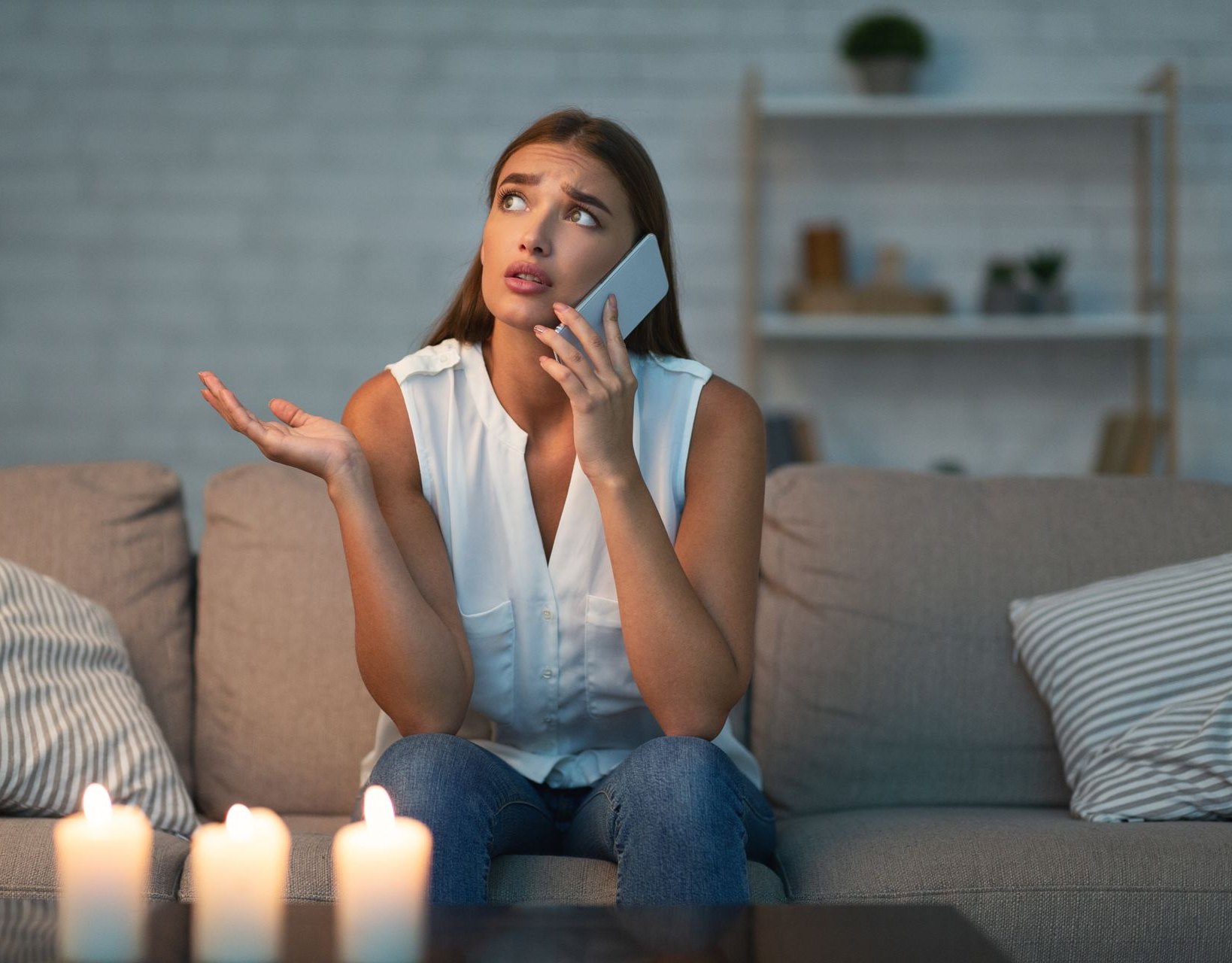 Une femme assise sur un canapé, parlant au téléphone, l'air soucieux, avec des bougies allumées au premier plan.