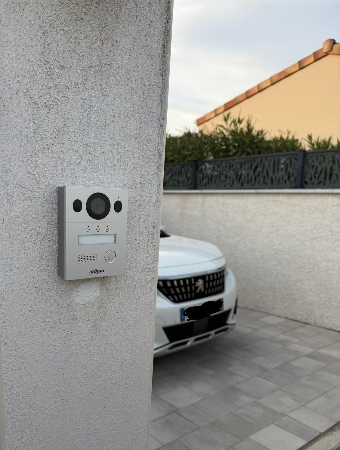 Interphone de portail d'entrée sur un mur en stuc, avec une voiture blanche garée dans une allée derrière.