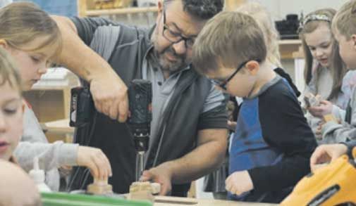 Ein Mann bringt einer Gruppe von Kindern in einem Klassenzimmer den Umgang mit einer Bohrmaschine bei.