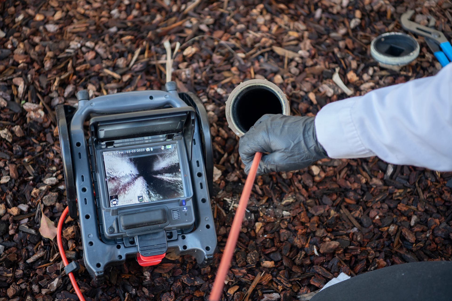 Un plombier inspecte une canalisation avec une caméra, entouré de paillis, tenant un câble.