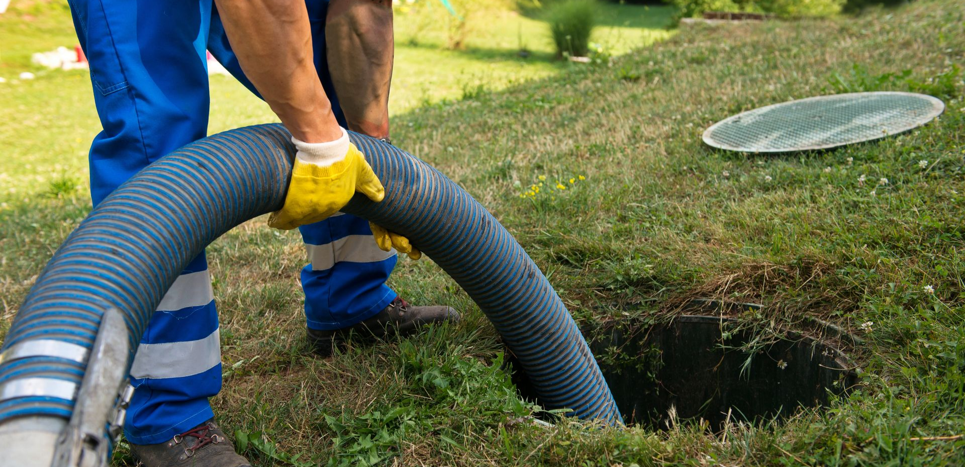Une personne en salopette bleue avec des gants jaunes tient un tuyau de vidange au-dessus d'une fosse septique ouverte.