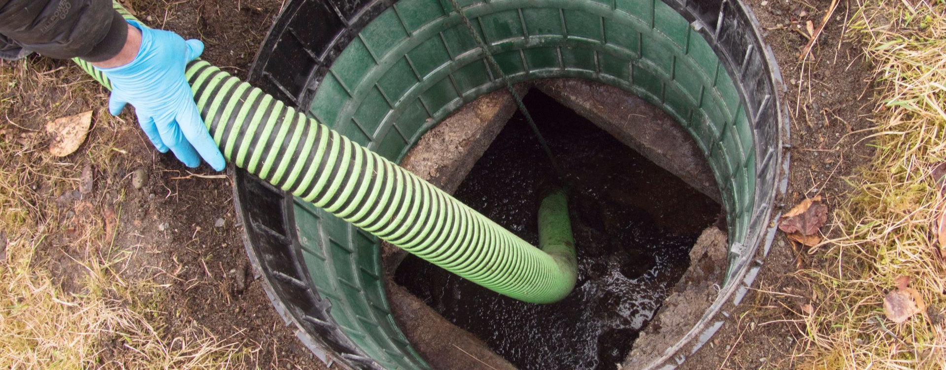 Une personne portant des gants bleus tient un tuyau et vide une fosse septique, dont l'ouverture est entourée d'un anneau vert.