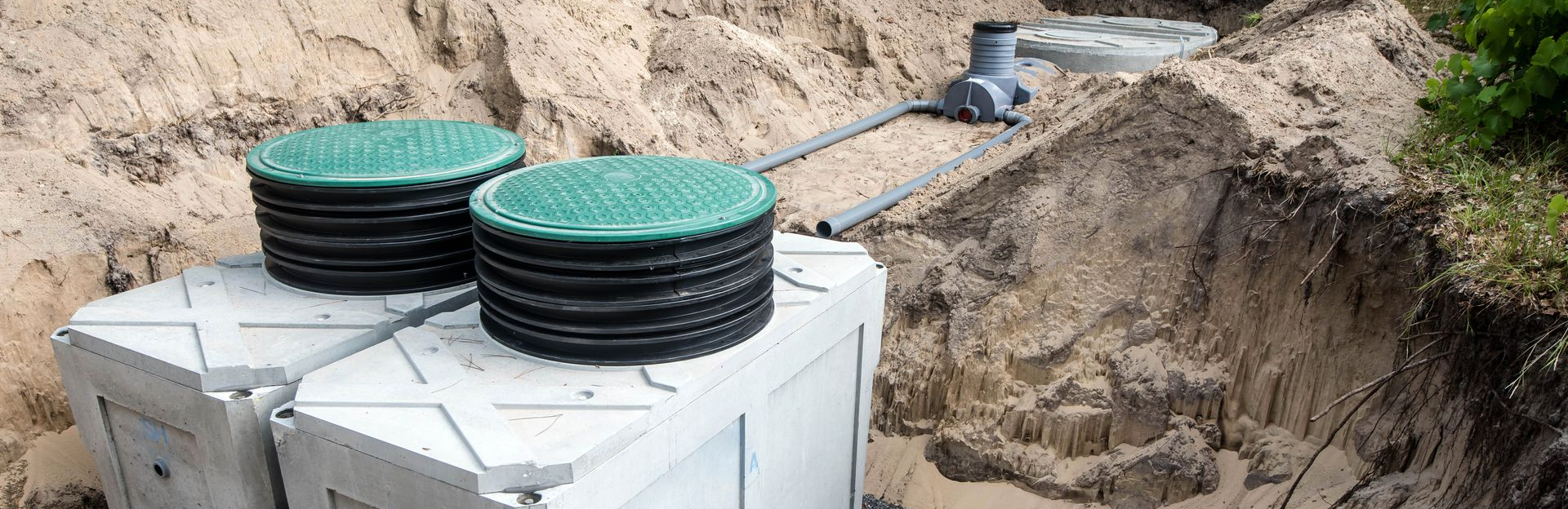 Deux couvercles de fosse septique, l'un noir et l'autre vert, sont posés au-dessus d'une cuve blanche dans une zone excavée.