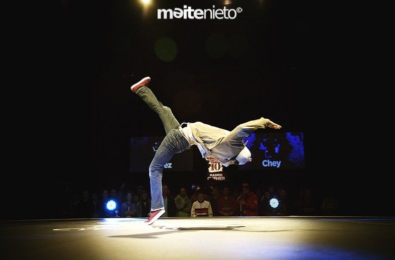 Un bailarín de breakdance realiza un movimiento en el escenario con luces y multitud de fondo.
