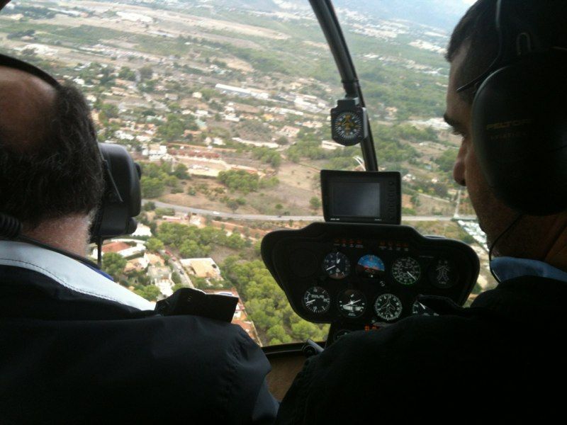 Dos personas en la cabina de un helicóptero, mirando un paisaje de edificios y árboles.