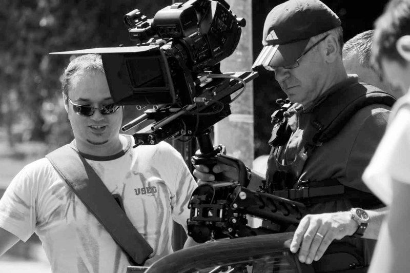 Dos hombres manejan una cámara de video en un día soleado. Uno ajusta el equipo, el otro observa.