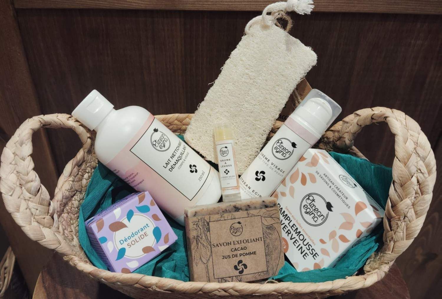Una cesta de mimbre llena de productos para el cuidado de la piel, que incluye una loción, jabones, un exfoliante y una esponja vegetal sobre un paño de color turquesa.