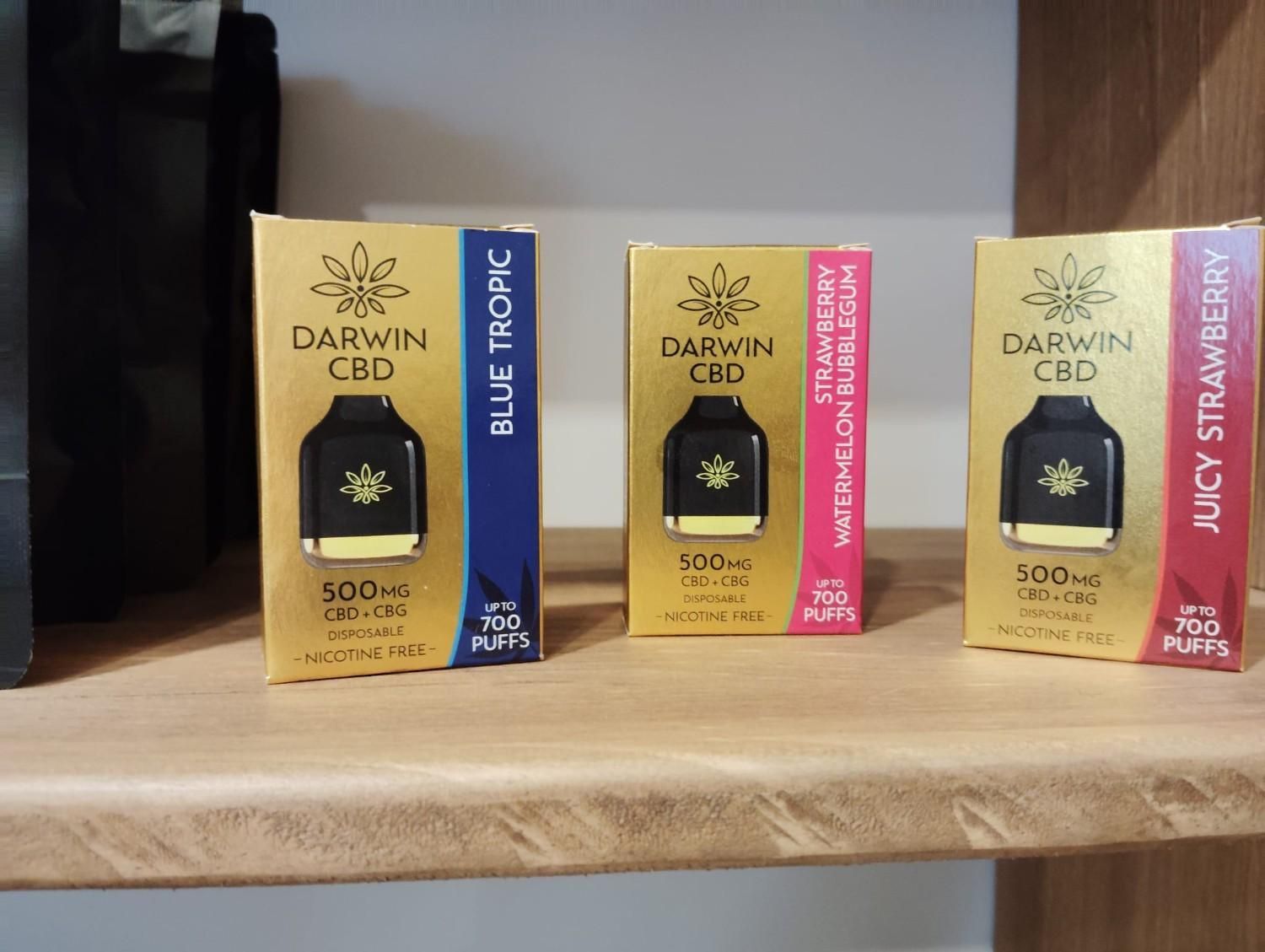 Tres cajas desechables para vaporizador Darwin CBD en los sabores Blue Tropic, Strawberry Watermelon y Juicy Strawberry reposan sobre un estante de madera.