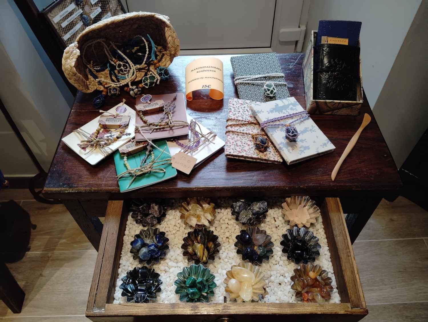 Una mesa de madera con objetos artesanales, entre los que se incluyen pequeñas libretas, joyas y un cajón lleno de cuencos con racimos de cristal.