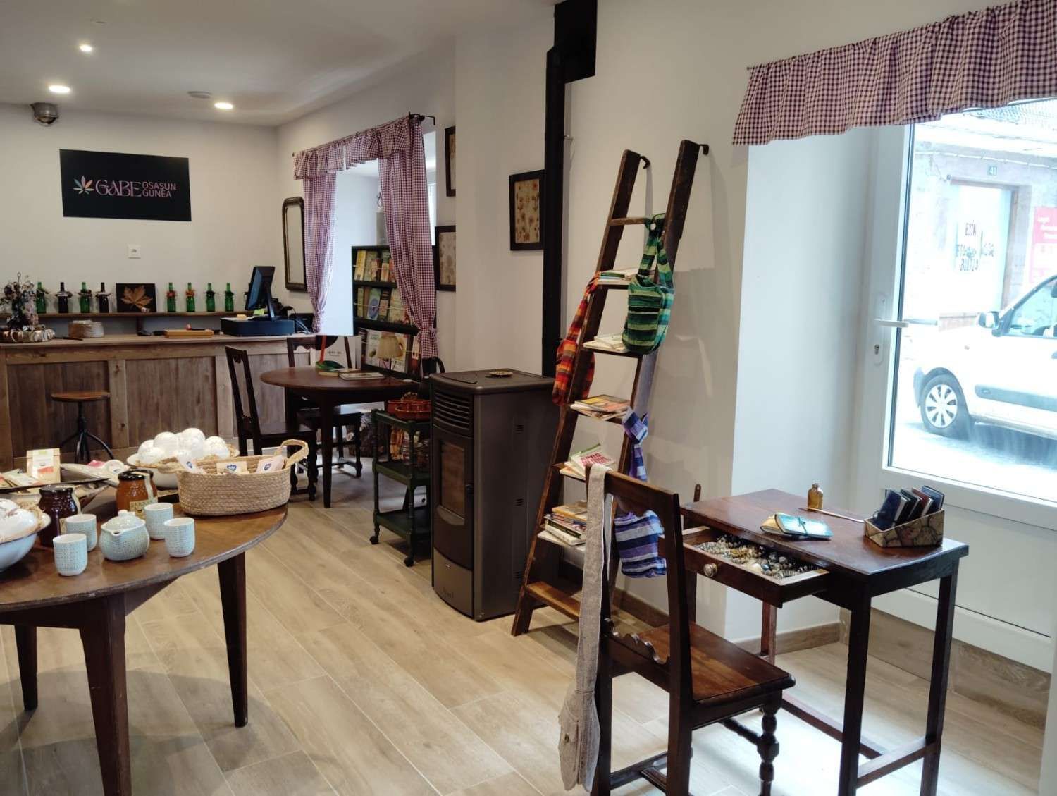 Un acogedor interior de tienda con mesas de madera, una estufa de pellets, una escalera de madera apoyada sobre la que se exhiben los artículos y una ventana con vistas.