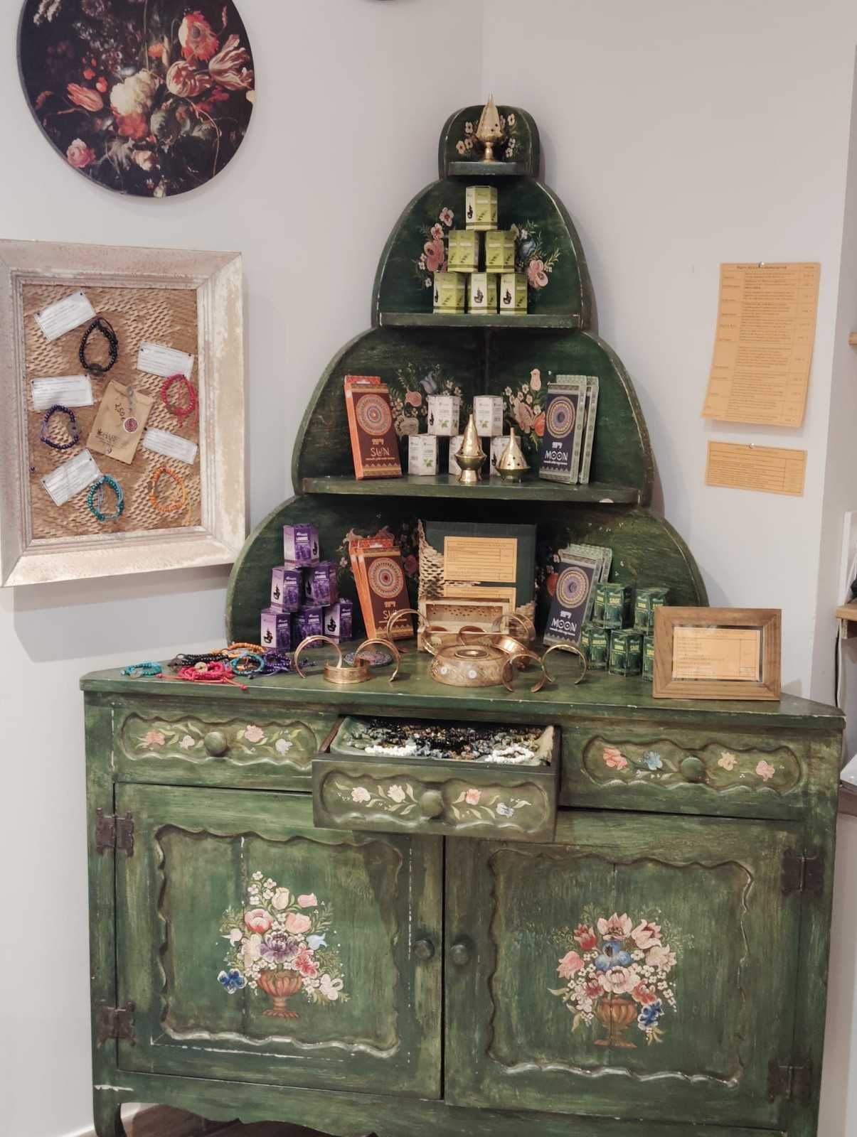 Una vitrina esquinera arqueada pintada de verde, que exhibe latas de té, libros y baratijas, con un tablón de corcho en la pared a su izquierda.