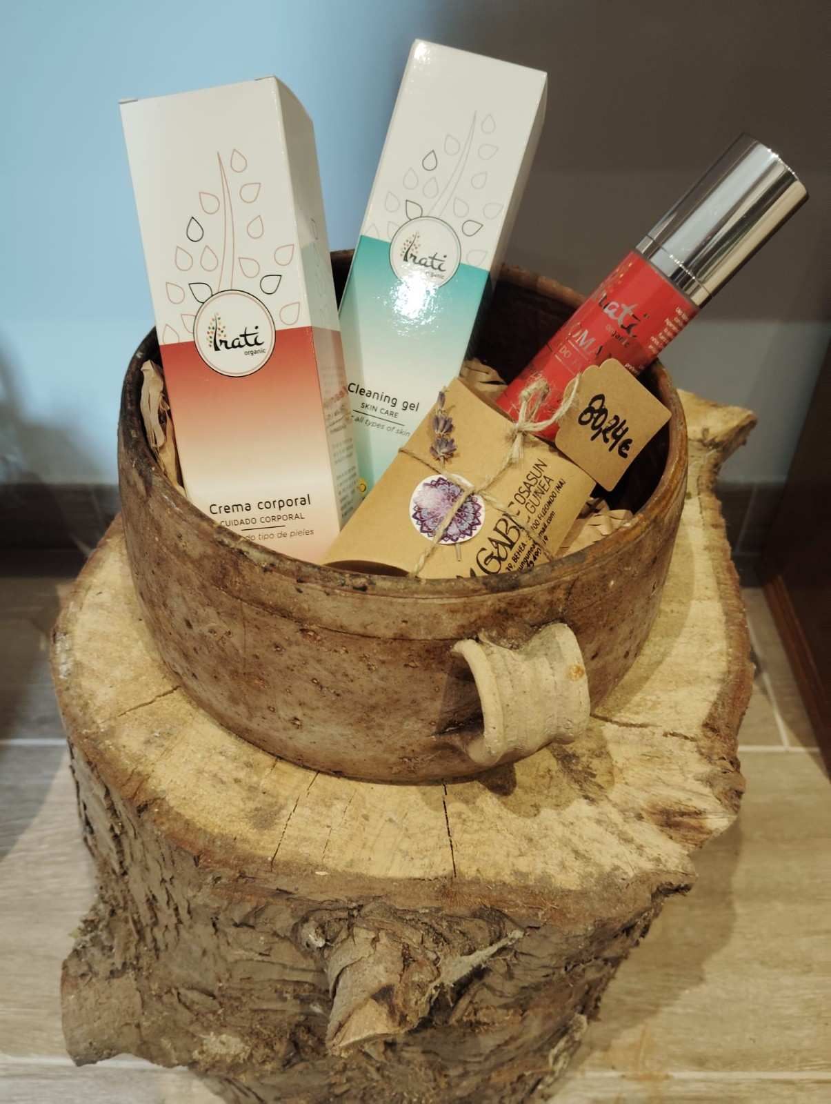 Dos cajas de cosméticos y un tubo rojo en una cesta rústica de madera, que descansa sobre un grueso pedestal hecho con un tocón de árbol.