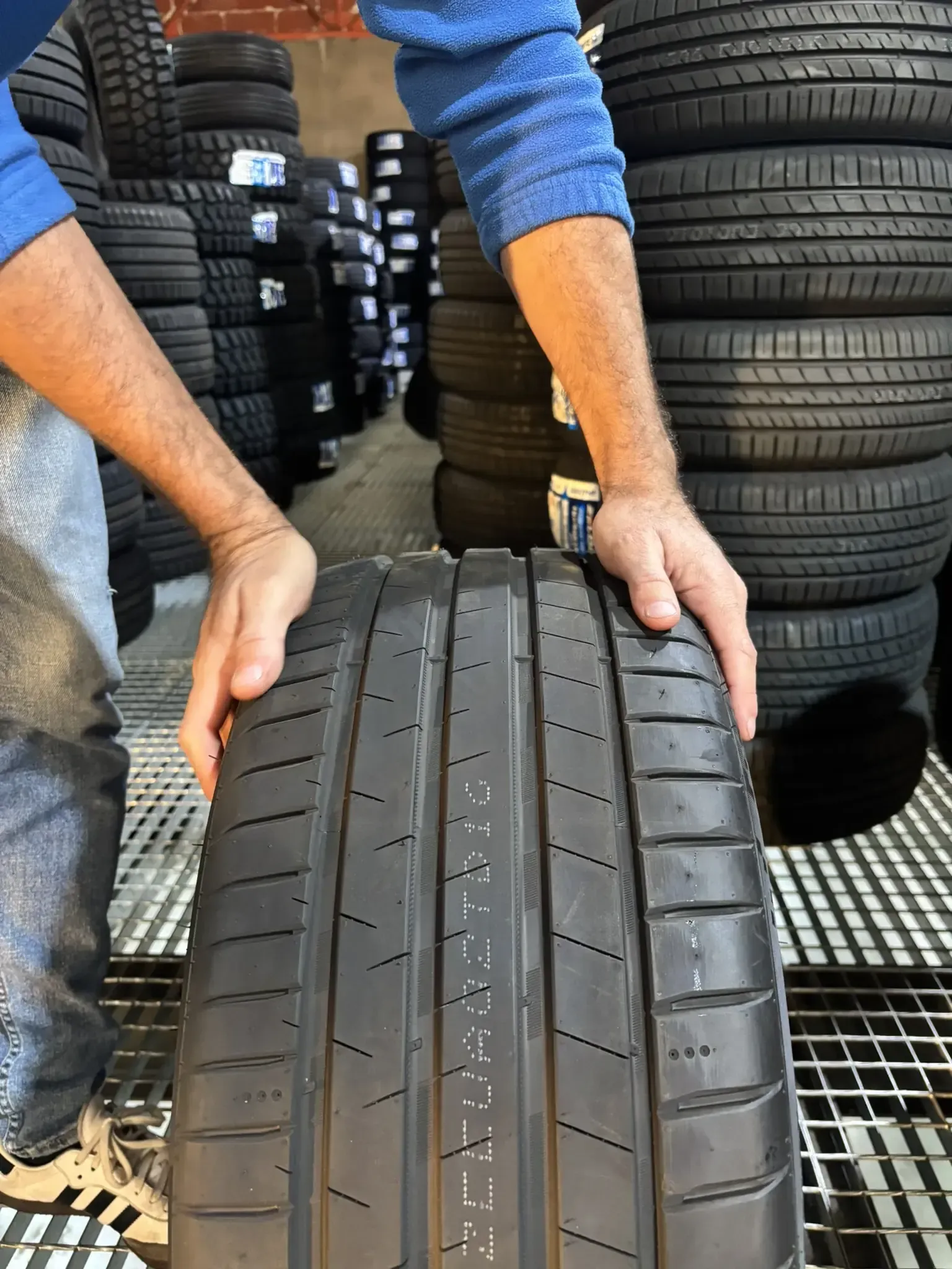 Persona sosteniendo un neumático en una tienda de neumáticos, con pilas de neumáticos en el fondo.