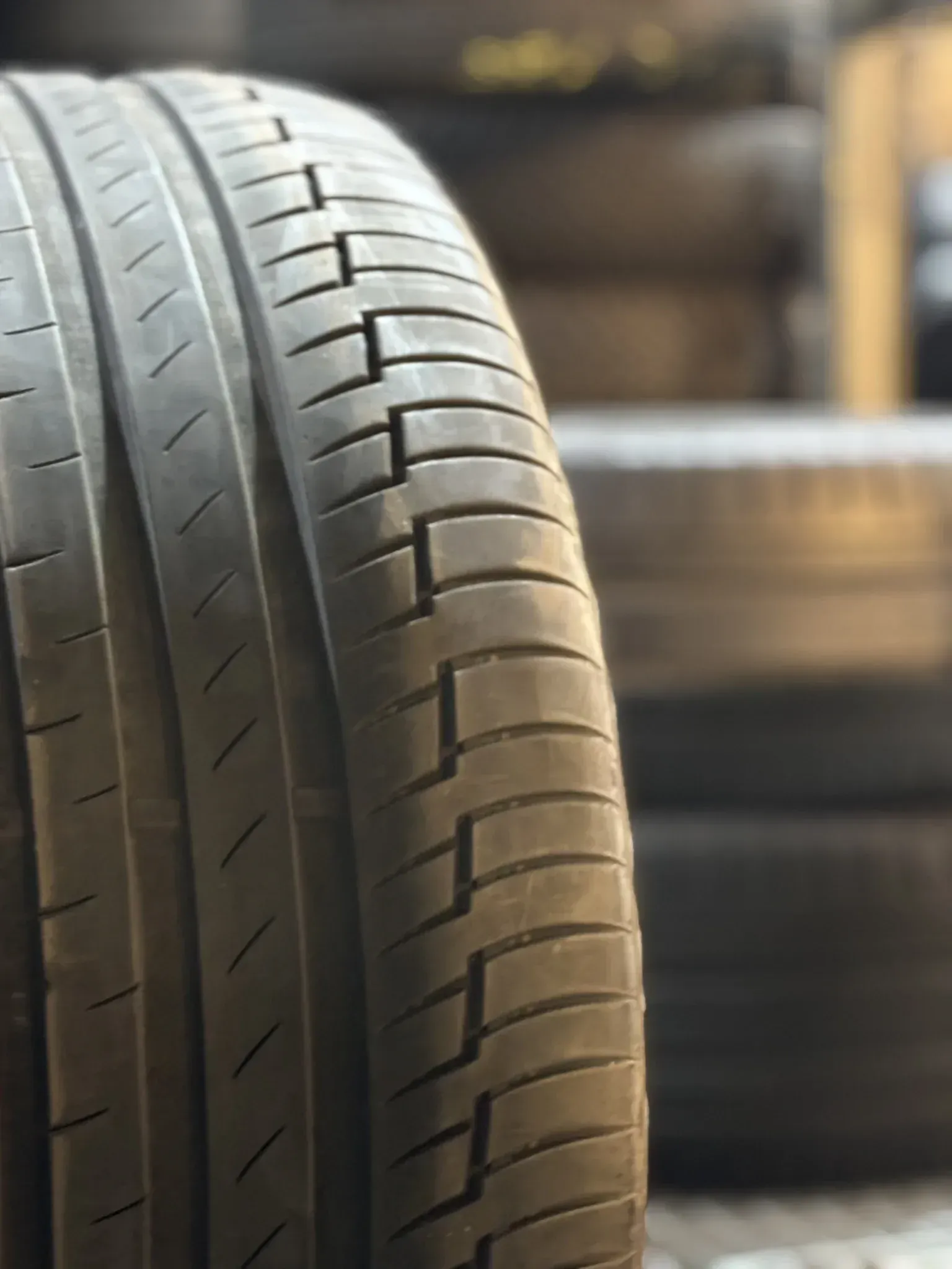 Primer plano de un neumático negro con dibujo de banda de rodadura, delante de una pila borrosa de neumáticos.