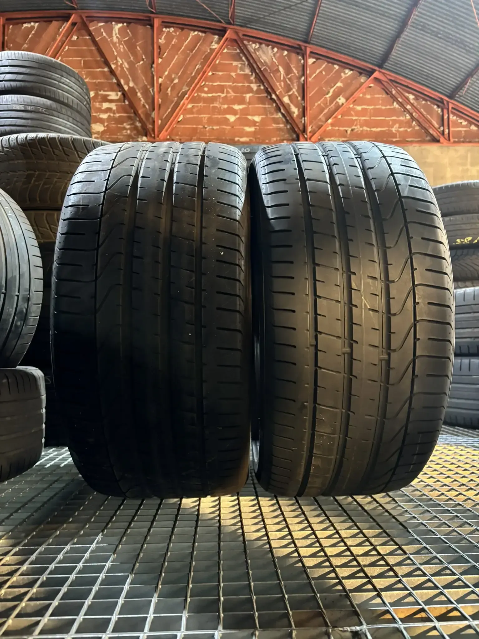 Dos neumáticos de automóvil negros uno al lado del otro, colocados sobre un soporte de metal, con otros neumáticos visibles en el fondo.