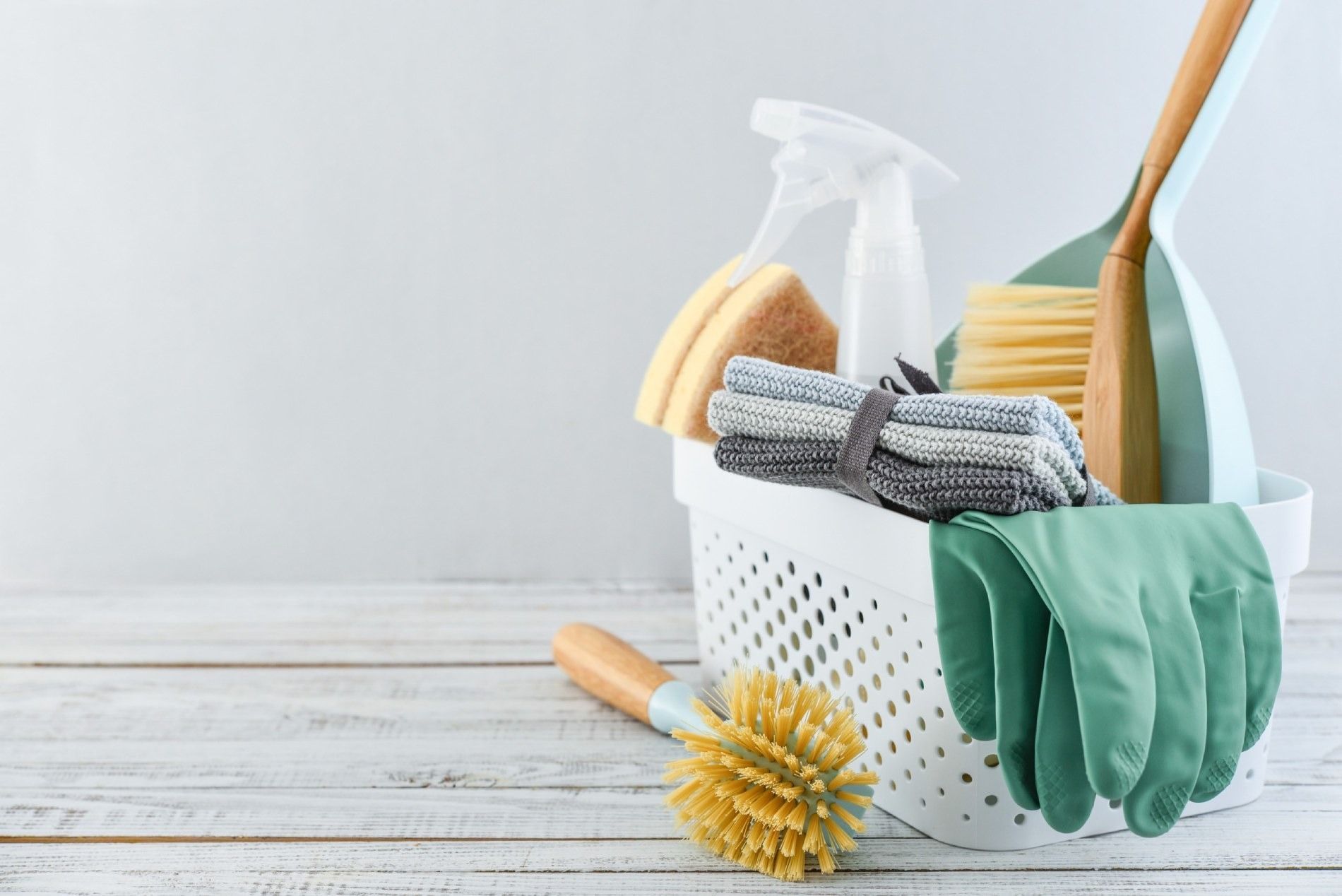 Artículos de limpieza en una cesta blanca: botella de spray, esponjas, toallas, cepillo, escoba y guantes.