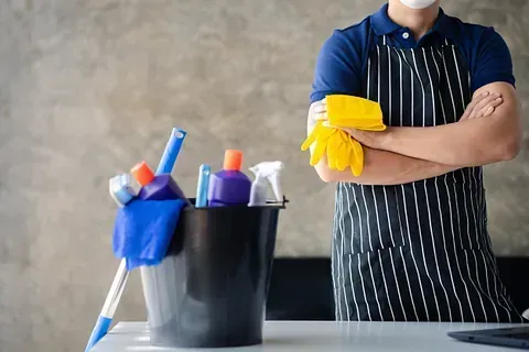 Persona con delantal y guantes, de pie junto a productos de limpieza en un balde.