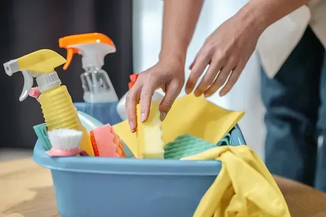 Persona que mete la mano en un balde azul lleno de productos de limpieza: botellas de spray, esponjas, cepillos y paños.