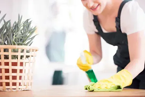Mujer con mono y guantes amarillos limpiando una superficie de madera con una botella de spray y un paño.