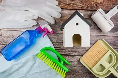Productos de limpieza dispuestos cerca de una pequeña maqueta de casa blanca sobre una superficie de madera.