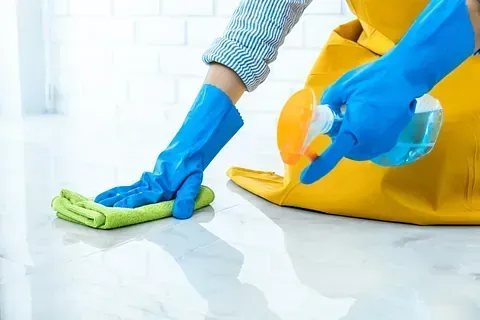 Persona limpiando piso con atomizador y paño, vistiendo guantes azules y delantal amarillo.